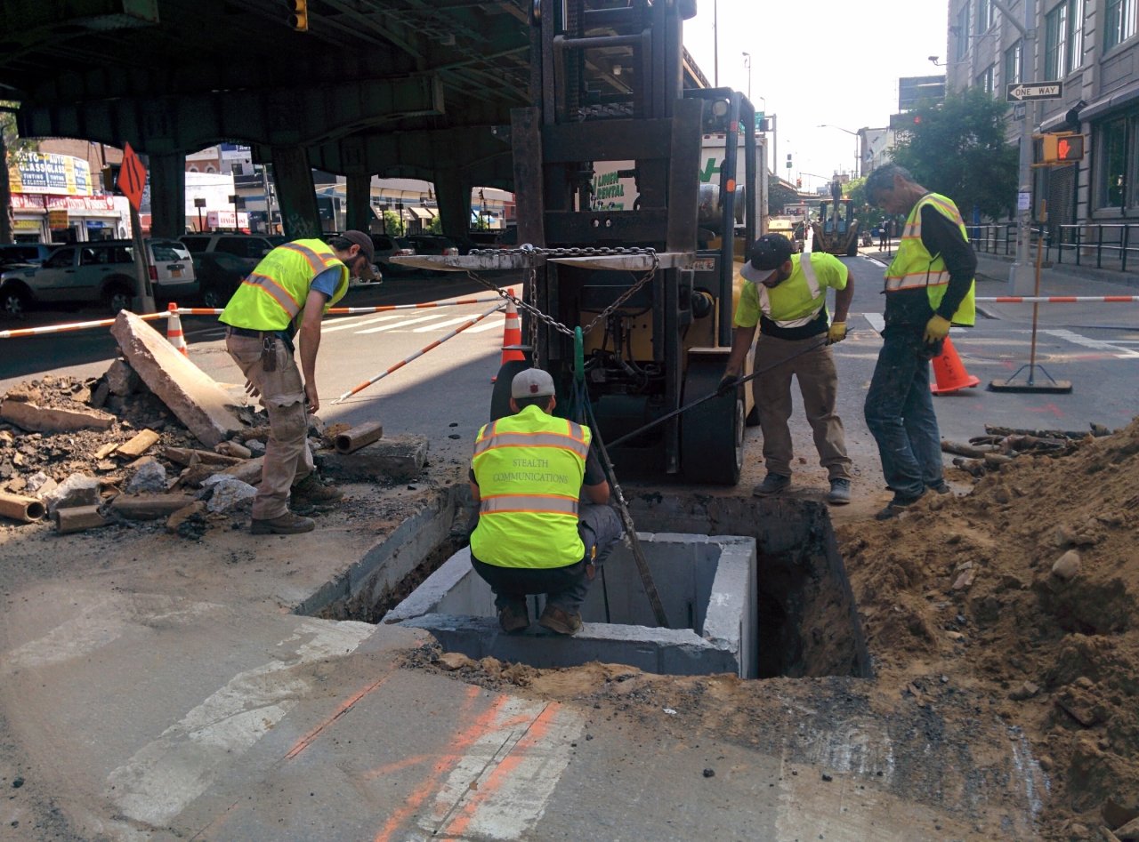 Stealth crew positioning a rectangular underground access chamber at a street construction site.