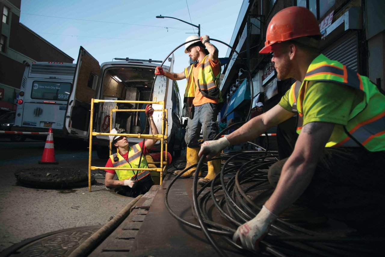 Stealth crew feeding cable into an underground access opening beside a service van.