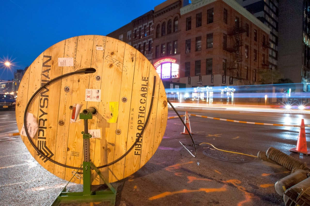 Large wooden fiber-optic cable reel beside a city street, with traffic light trails in the background at dusk.